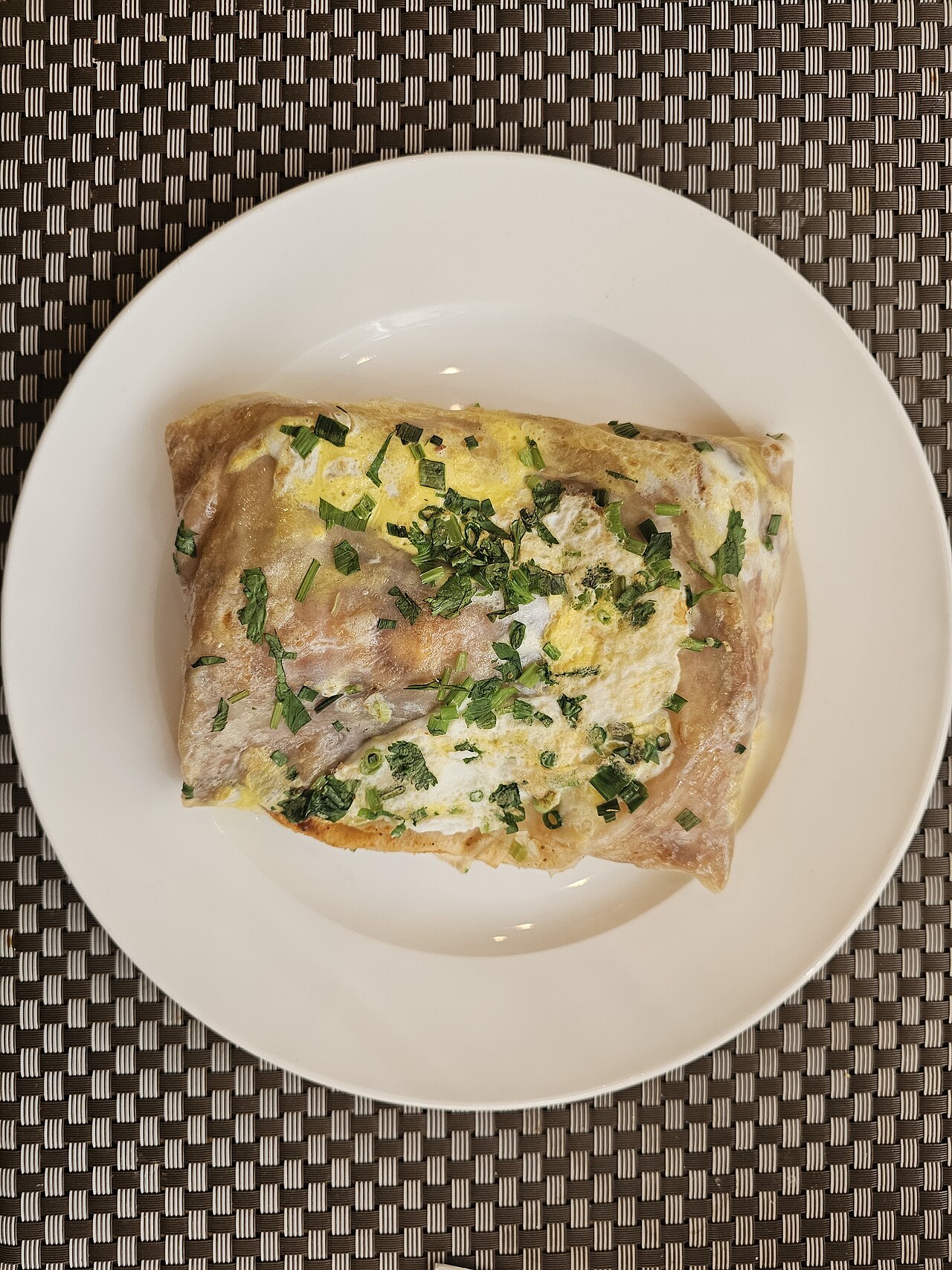 A jianbing — a folded egg crêpe parcel scattered with scallions and coriander, on a white plate