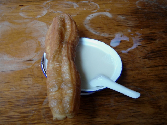 A golden crispy youtiao (fried dough stick) next to a bowl of hot soy milk with a spoon, on a wooden table — the classic Chinese breakfast pairing
