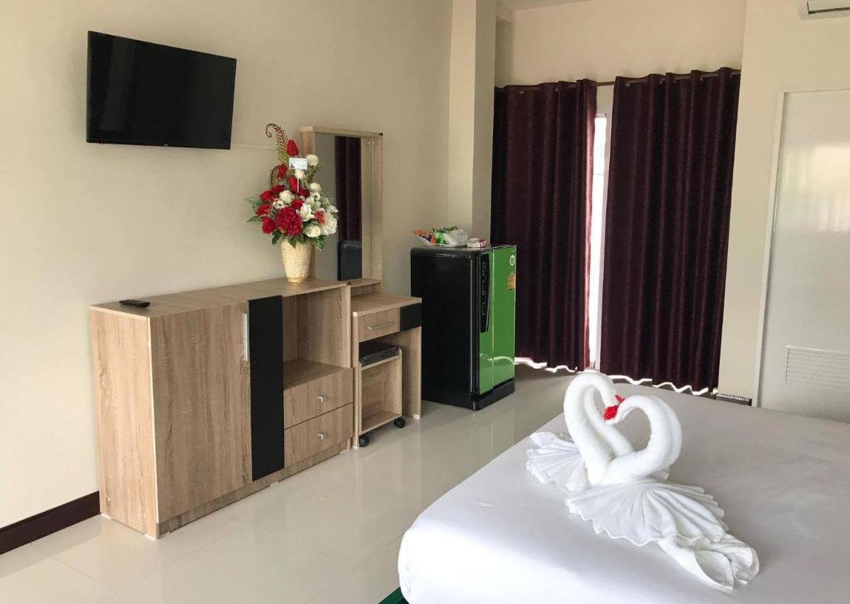 ห้องพักมุมโต๊ะเครื่องแป้ง ตู้เย็นเขียว ทีวีติดผนัง และผ้าเช็ดตัวพับเป็นรูปหงส์บนเตียง