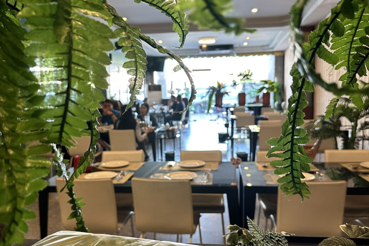 ห้องอาหารของรีสอร์ท มองผ่านเฟิร์นเขียวที่แขวนประดับ มีโต๊ะยาวจัดจานเรียบร้อย