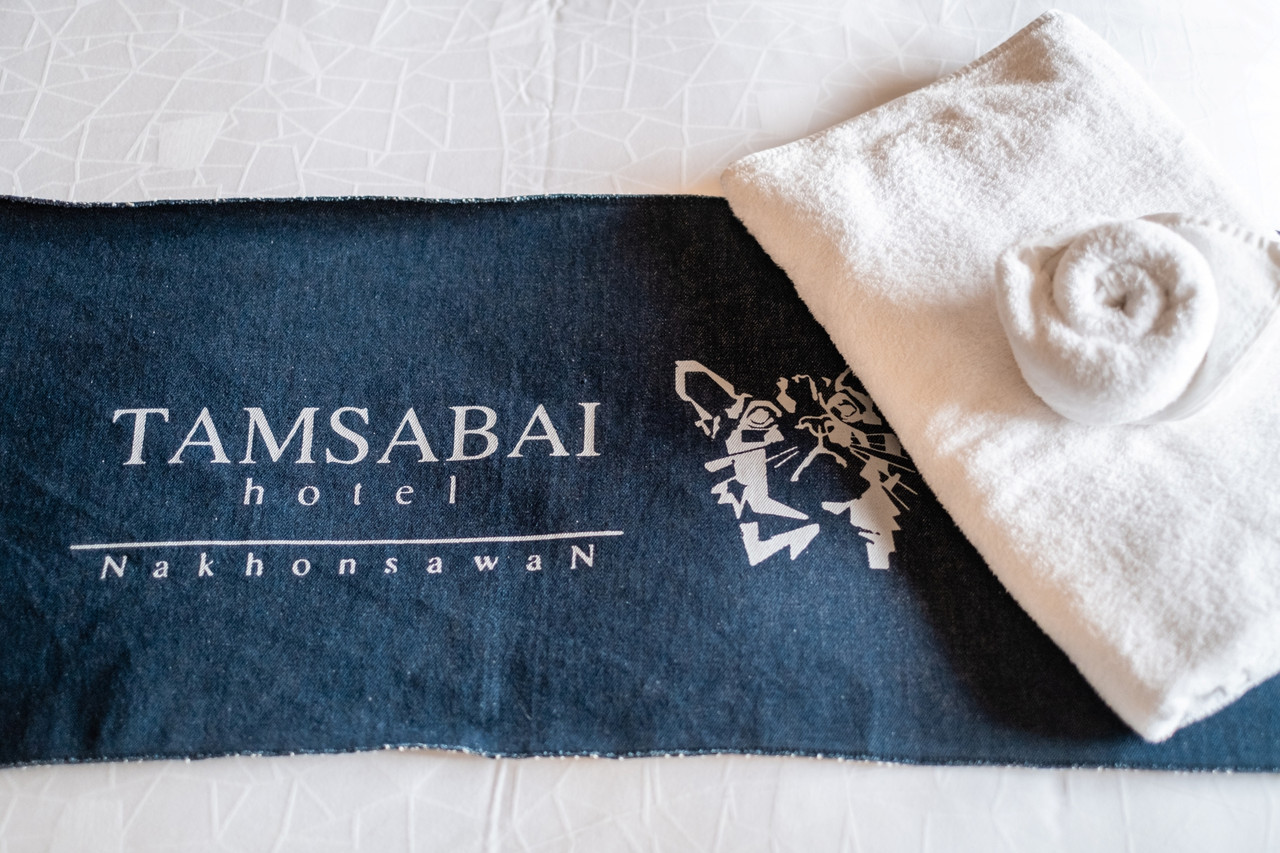 ผ้าเช็ดตัวสีกรมท่าพิมพ์โลโก้ TAMSABAI hotel Nakhonsawan กับรูปแมว วางบนเตียงผ้าขาว