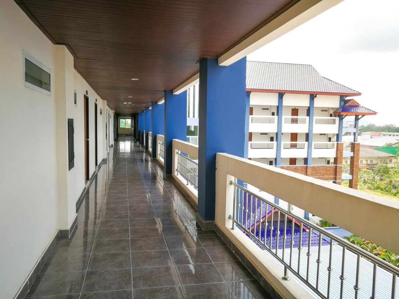 ระเบียงทางเดินชั้นบนของ Din Khao Apartment พื้นกระเบื้องมันวาว ราวเหล็ก เสาสีน้ำเงิน มองเห็นอีกปีกของตึกและวิวเมืองนราธิวาส