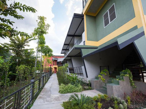 Two-storey green-and-grey building with yellow structural columns and a concrete path through the planted garden