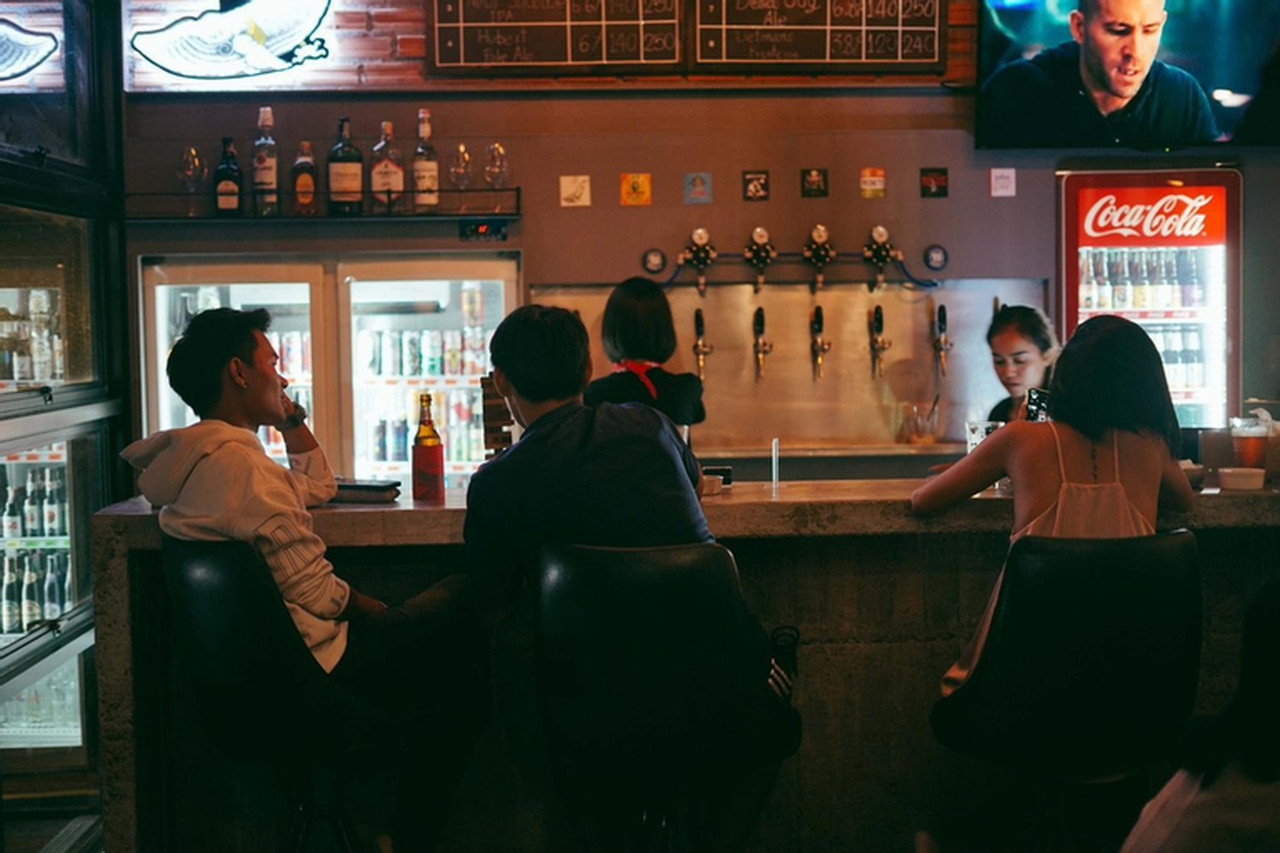 บาร์ของ Small Town Hostel พัทลุงตอนกลางคืน แขกนั่งที่เคาน์เตอร์ปูน มีตู้แช่เครื่องดื่ม หัวจ่ายเบียร์สด และจอทีวีเหนือบาร์ บรรยากาศแสงสลัวอบอุ่น