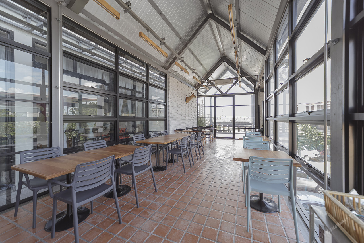 มุมคาเฟ่-ร้านอาหารชั้นบนของ Small Town Hostel พัทลุง โต๊ะไม้เก้าอี้สีเทาเรียงริมกระจกบานใหญ่ หลังคาทรงจั่วโครงเหล็ก พื้นกระเบื้องดินเผา แสงธรรมชาติส่องเข้า