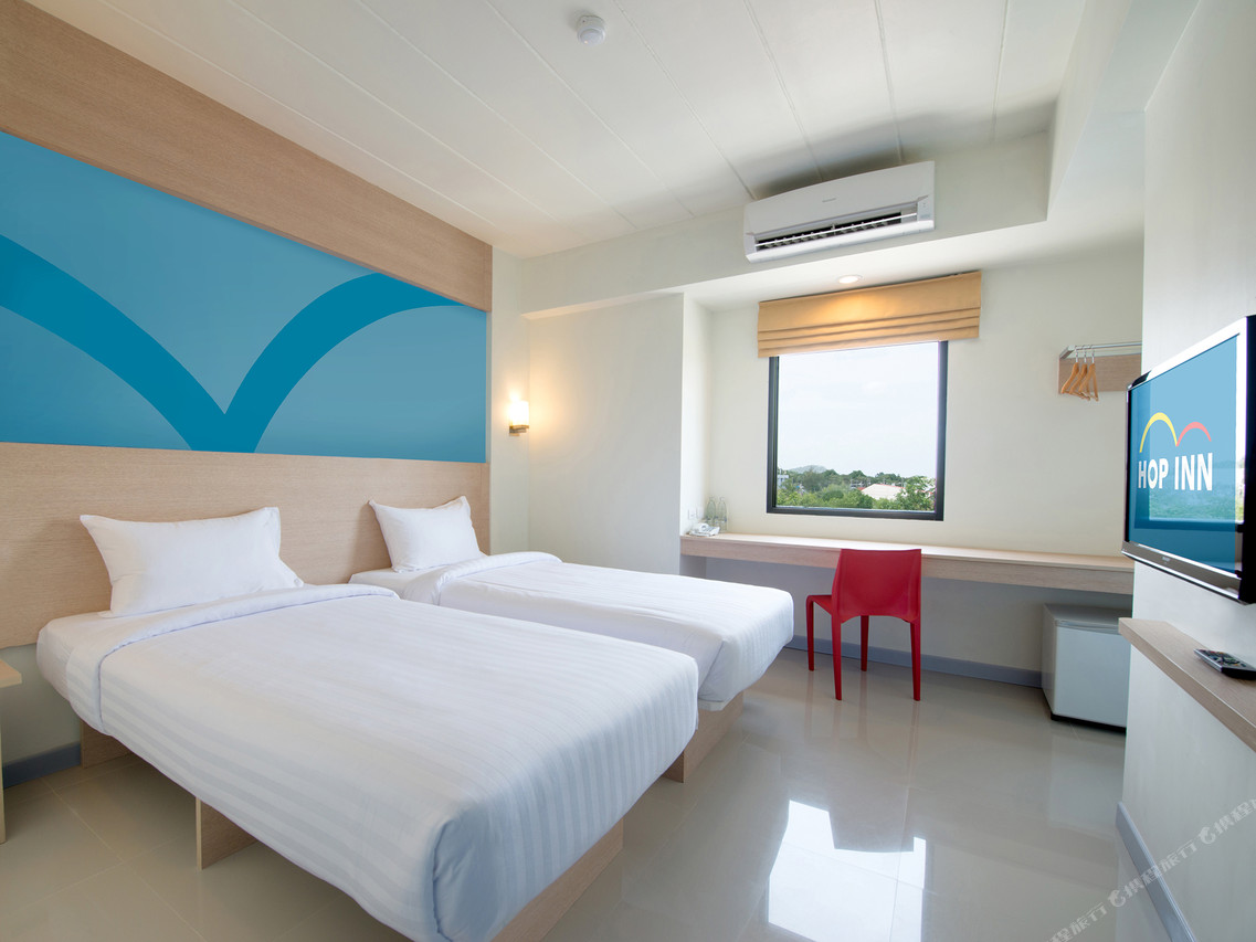 Standard Twin room at Hop Inn Phetchabun with two single beds, the brand's curved blue headboard graphic, a red plastic chair, a long window desk and a wall-mounted TV showing the HOP INN logo