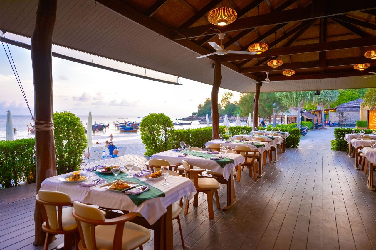 Open-air beach restaurant looking out to the sand and longtail boats at sunset