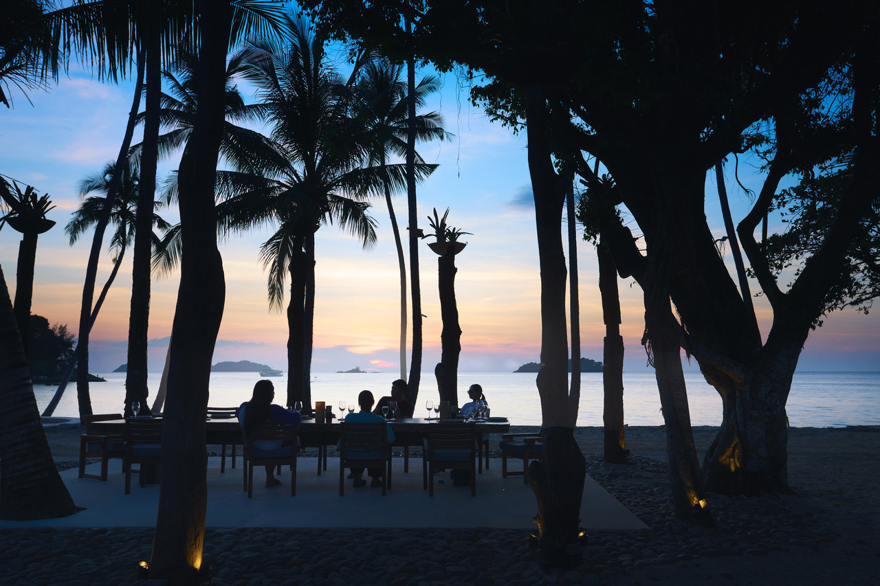 Beachfront dining table on the sand under coconut palms with a sunset over the sea at Koh Chang