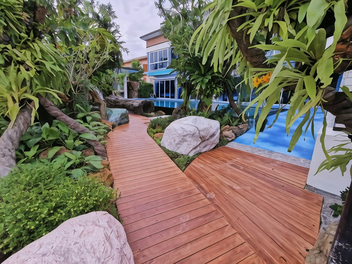 A winding wooden boardwalk through the garden with large boulders, looking toward the building and pool at Uthaithani Resort
