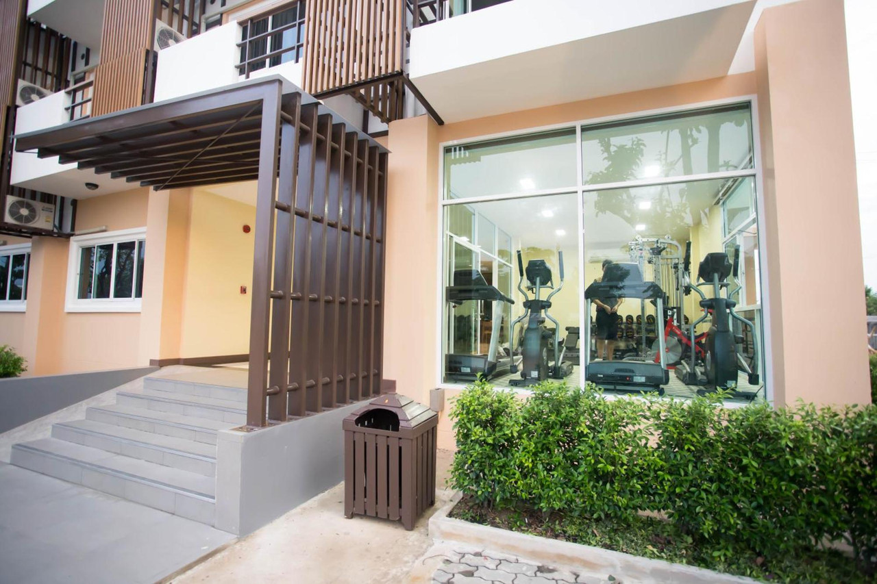 ทางเข้าอาคารและห้องฟิตเนสกระจกของ Thaimit Resort มองเห็นลู่วิ่งและเครื่องออกกำลังกายด้านใน บันไดทางขึ้นและพุ่มไม้